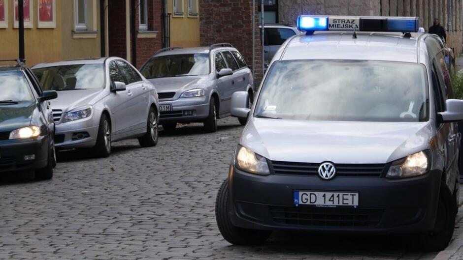 radiowóz straży miejskiej zaparkowane samochody wybrukowana jednia pieszy fragmenty budynków