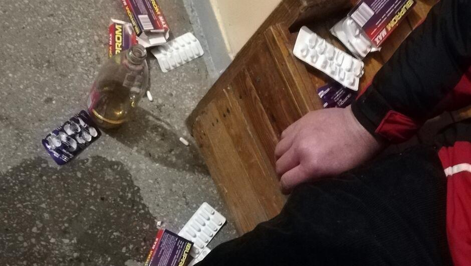 opakowania po tabletkach Ibuprom otwarta buteka wina rozrzucone na podłodze z lastryko fragment ławka fragment siedzącego mężczyzny