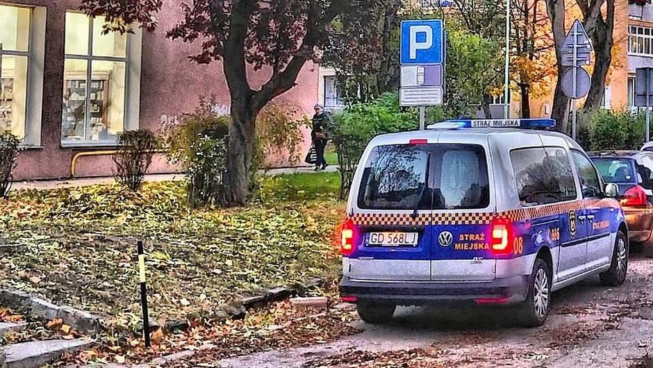 radiowóz zaparkowany samochód osobowy znaki drogowe drzewa sklep trawnik pokryty liśćmi blok mieszkalny