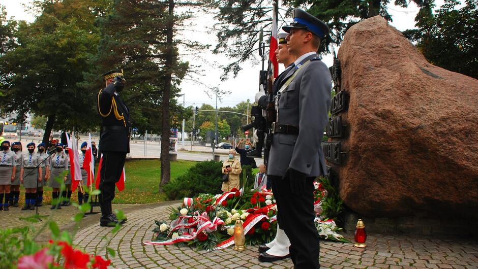 strażnik oddaje honor przed pomnikiem warta honorowa kwiaty znicze harcerze flagi Polski samochody ulica autobus drzewa.JPG