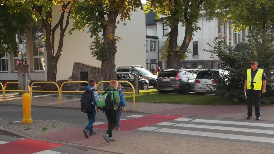 strażnik miejski pilnuje dzieci na przejściu dla pieszych w tle samochody i budynek szkoły.JPG