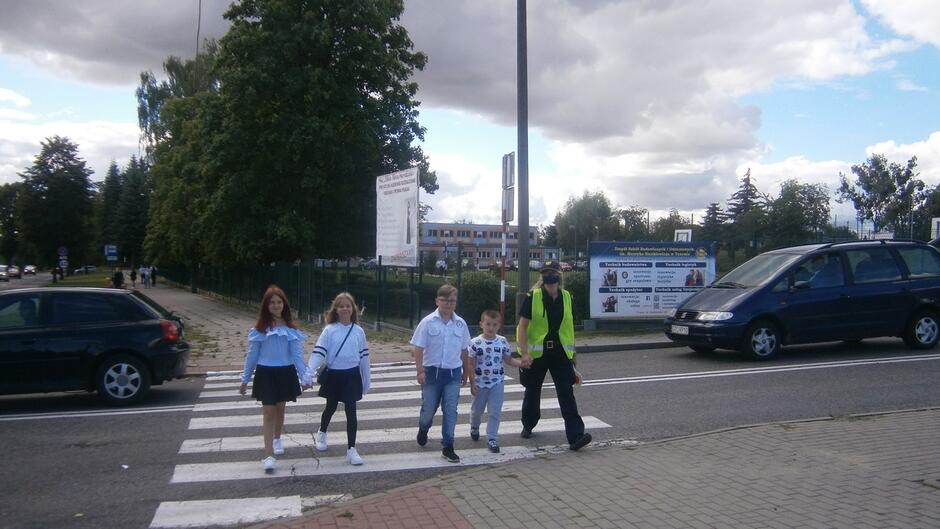 grupa dzieci wraz ze strażniczką miejską przechodzą przez przejście dla pieszych samochody budynek szkoły drzewa.JPG